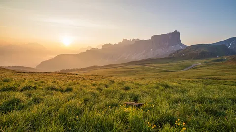 A green meadow with yellow flowers in front, a winding road and a steep mountain peak backlit by the sun.